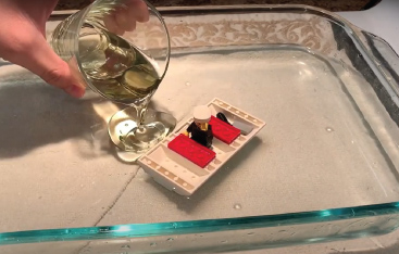 Close-up of a hand pouring oil into a dish of water containing a boat made of LEGO blocks.
