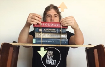 An educator holds "up" and "down" arrows in front of a stack of books sitting on a wooden board, balanced across the backs of two chairs.