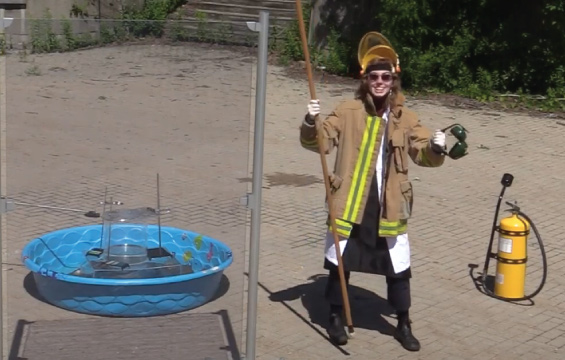 An educator in heavy protective gear prepares a combustible experiment outdoors in a kiddie pool.