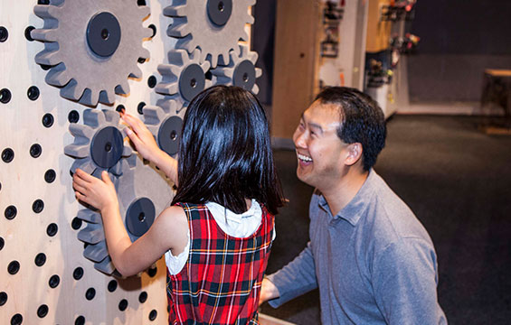 A parent looks on as a child explores giant gears.