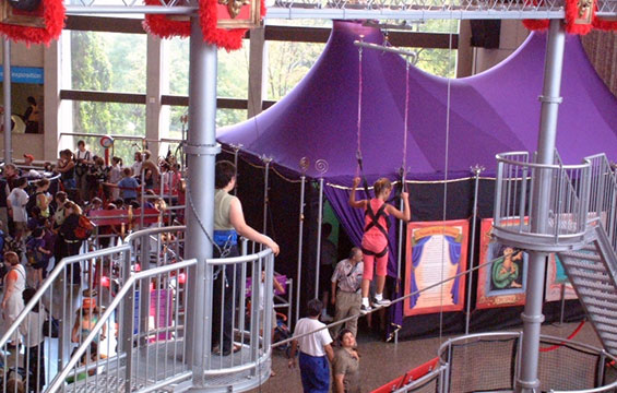 A person walks the high wire in front of large circus inspired tents.
