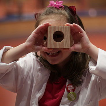 A young girl looks through a hole in a wood block.