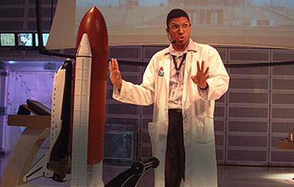 A host gives a presentation beside a large model of a space shuttle.