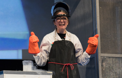 An educator wearing full safety gear gives two thumbs up, standing next to a smoking beaker