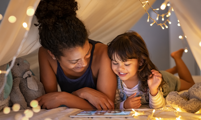 A smiling woman and child in an indoor blanket tent look at a tablet together.