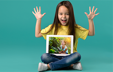 A young child holds sits cross-legged with a tablet in her lap, smiling and holding up her hands.