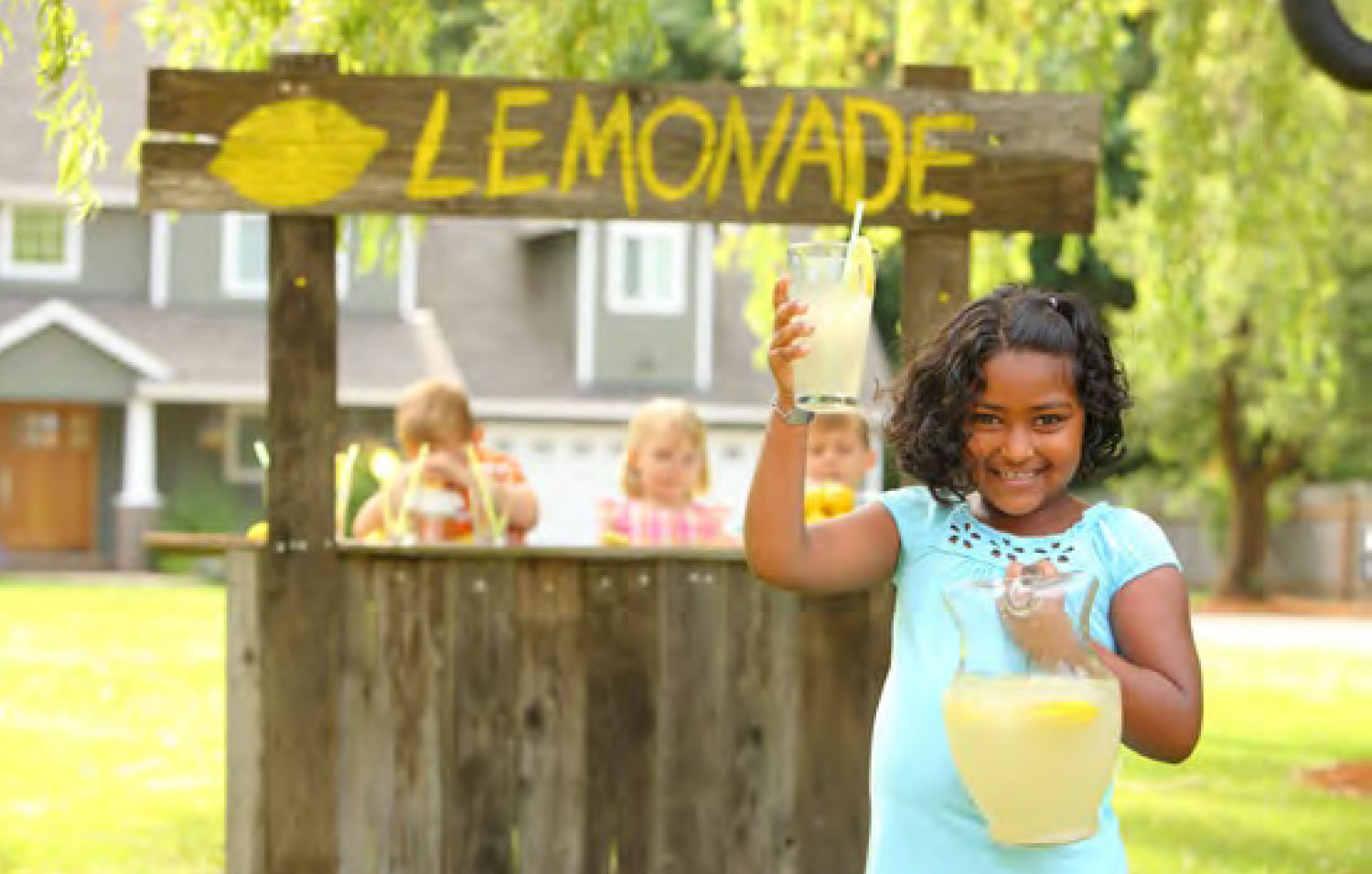 A girl holding a pitcher of lemonade in front of a lemonade stand.