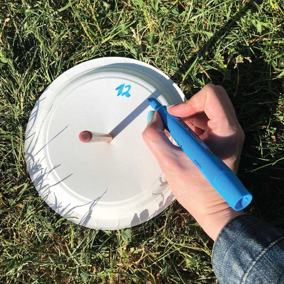 An upright pencil poked into the centre of a paper sits on the grass. Using a highlighter, a hand begins to draw numbers around the plate so it looks like a clock.