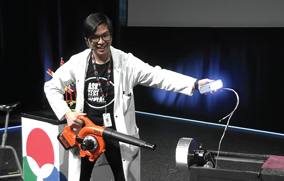 A Science Educator wearing a lab coat presents a demonstration with leaf blower in one hand and a glowing light in the other.