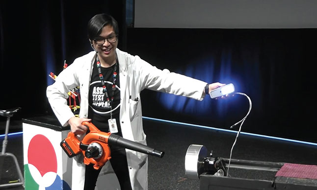 A Science Educator wearing a lab coat presents a demonstration with leaf blower in one hand and a glowing light in the other.