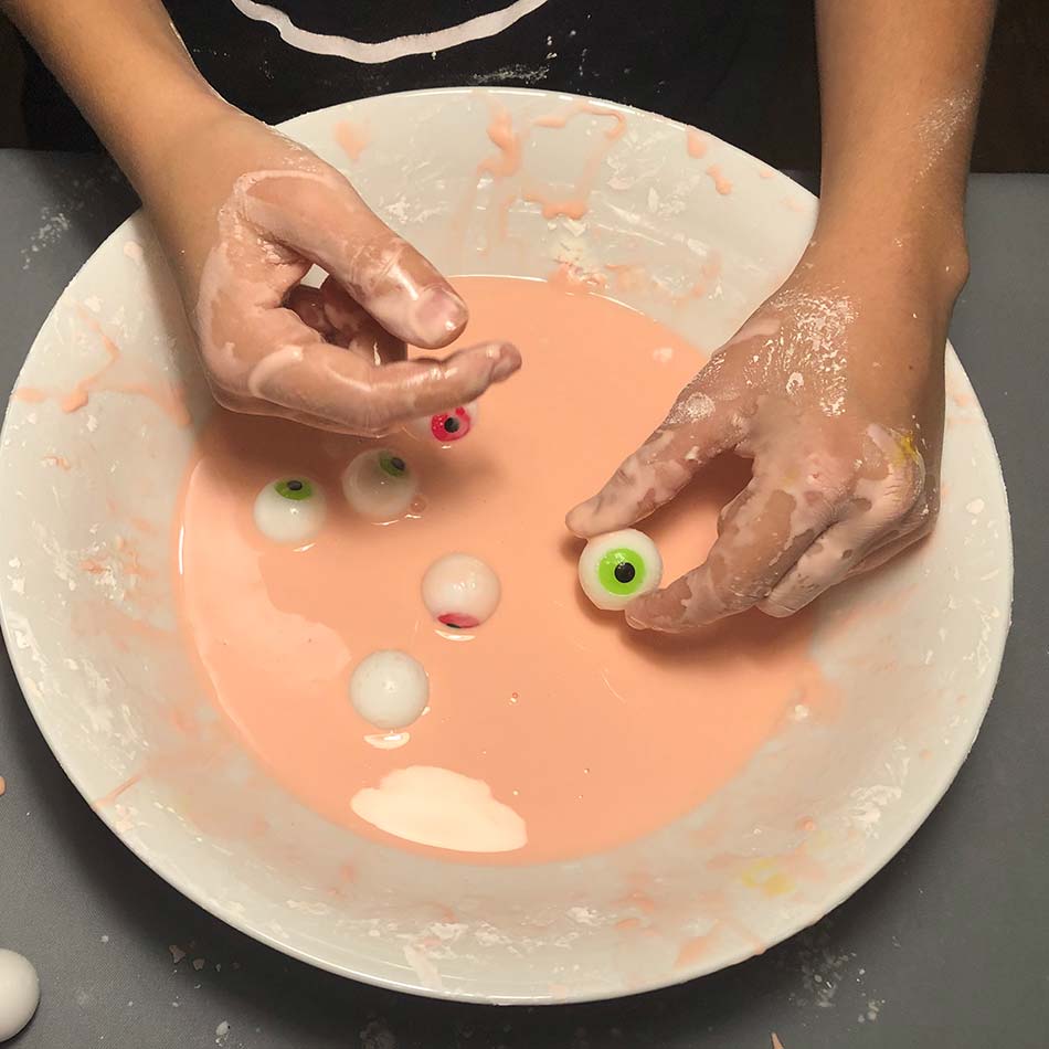 A child's hand holds a toy eyeball in a dish containing a pink slimy substance.
