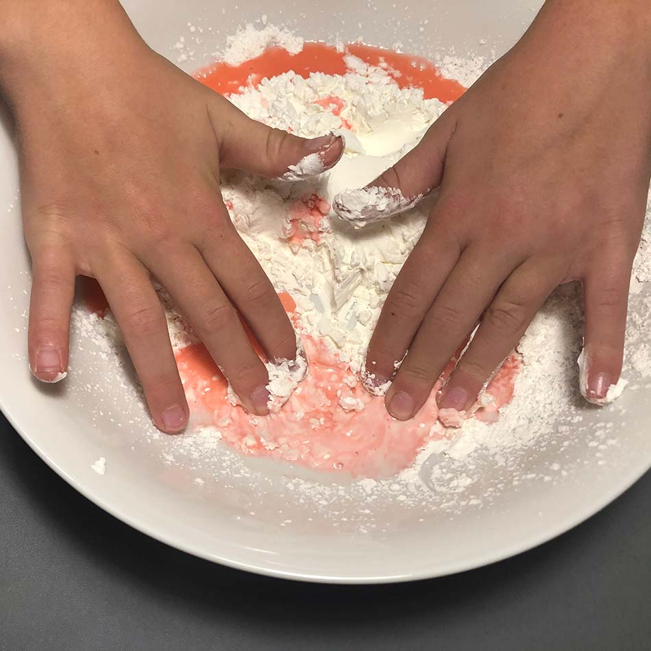 A close-up of child's hand mixing a red liquid into cornstarch on a plate.
