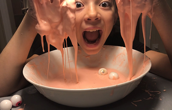 A smiling child hovers over a dish of pink Oobleck. Their hands are covered with Oobleck, which drips down into the dish.