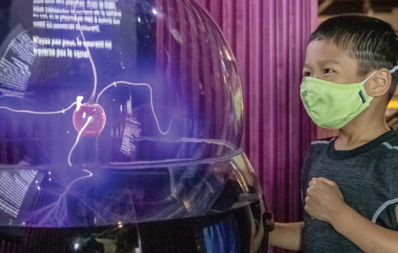 A child wearing a mask looks at a plasma ball.