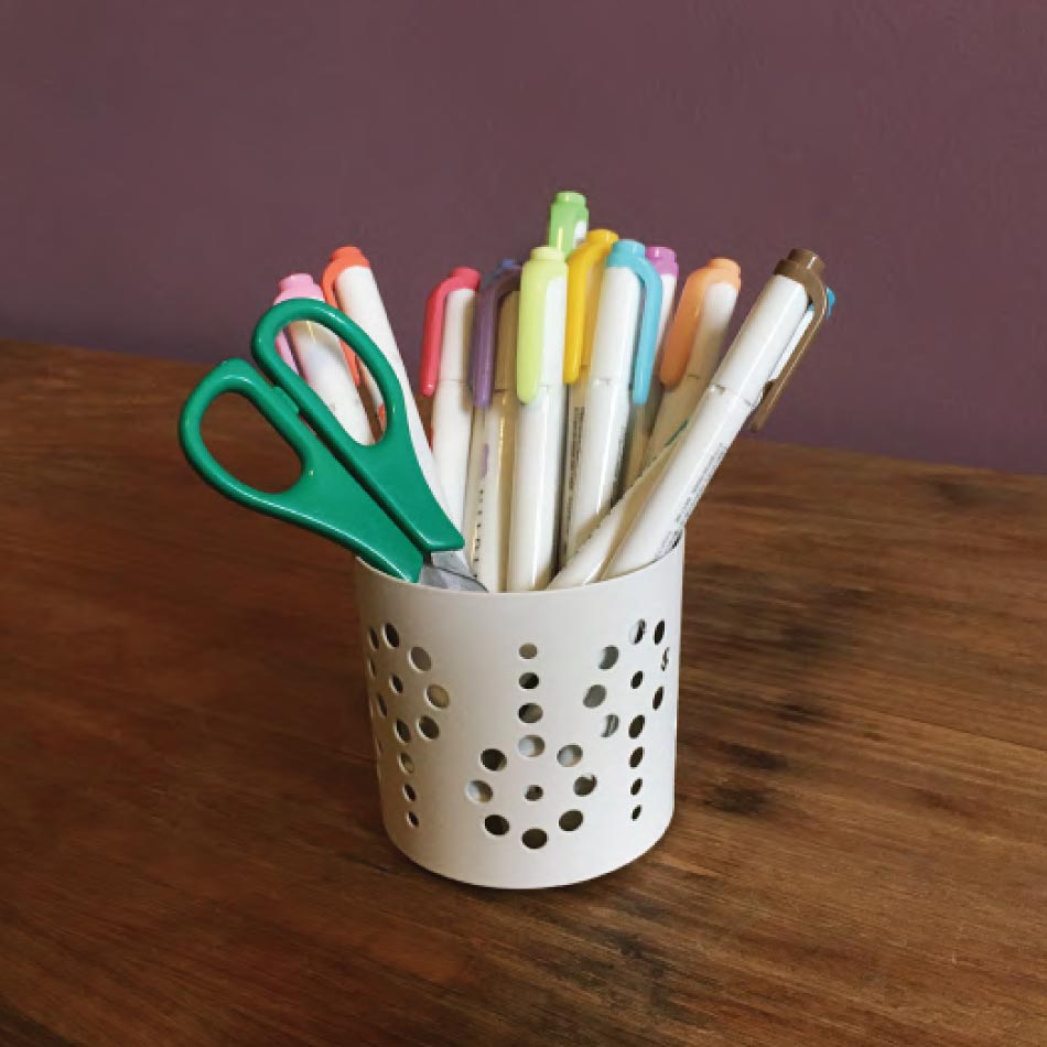 Photo of a jar sitting on a table containing a pair of scissors and several markers.