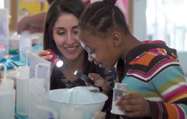 A teacher signs a flashlight into a bowl that a student peers into through a magnifying glass.