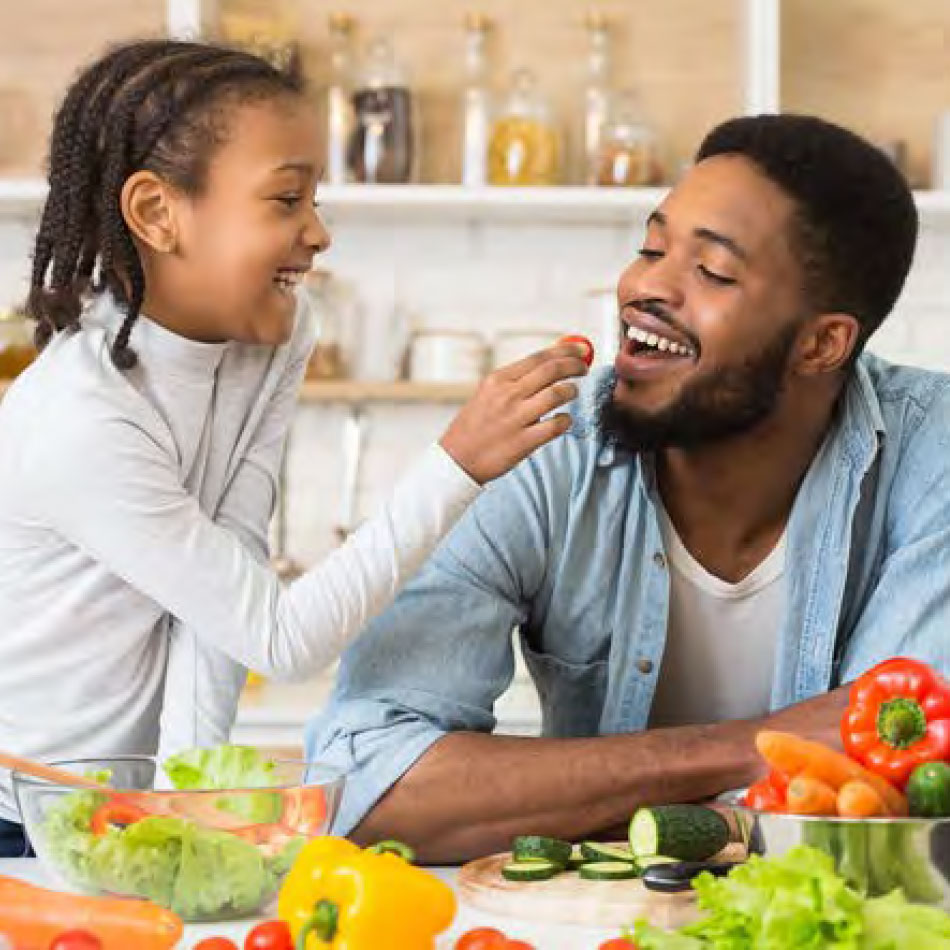 A girl feeding a tomato to a man with a salad being prepared.