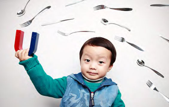 A boy holds a magnet while utensils fly comically towards it.
