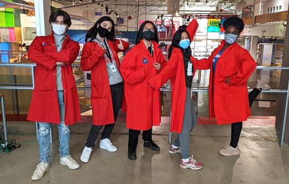 A group of Science School students wearing red lab coats pose in front of an exhibit hall.