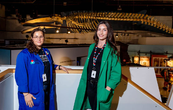 Two women in lab coats standing in front of a fin whale skeleton.