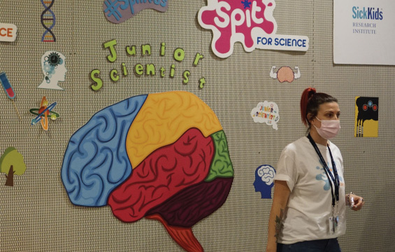 A SickKids researcher stands in front of a wall covered in colourful human brain illustrations and other science posters.