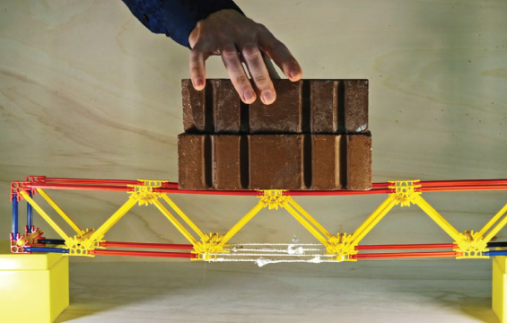 A hand balances two bricks on a bridge made out of a building toy.