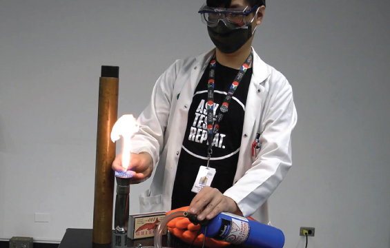An educator wearing goggles and a mask ignites a burner