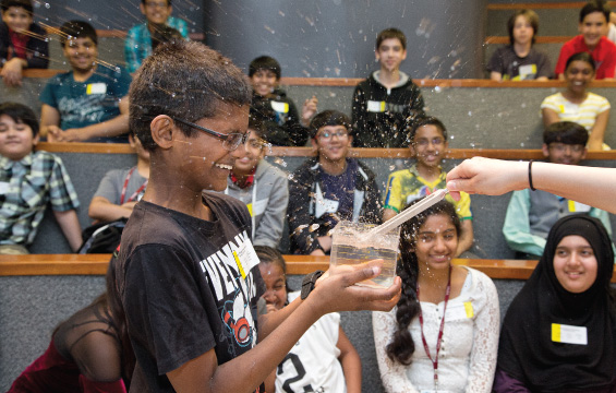 A vibrating tuning fork splashes water in a transparent vessel being held by a student.