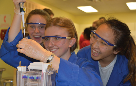 A student wearing a lab coat and safety goggles uses a pipette while two other students in safety equipment observe the process.