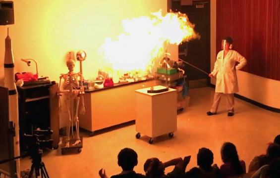 An educator wearing a safety mask initiates a spectacular combustion while students cover their ears.