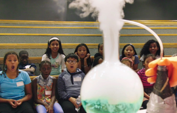 Students on benches react with amazement to a smoking beaker.