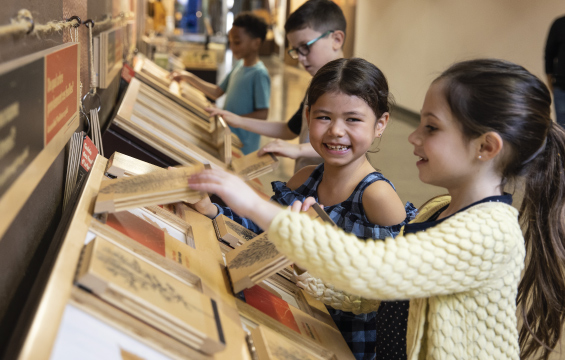 Smiling young students interact with an exhibit, lifting up wooden panels to read the information hidden underneath.