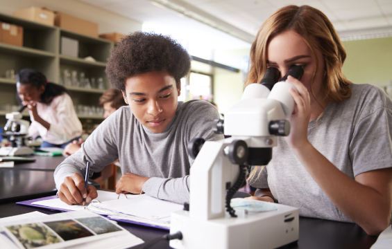 Two students work at a desk, with one peering into a microscope and the other writing in a binder.