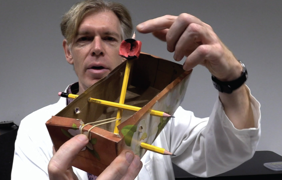 An educator demonstrates a catapult made out of a tissue box, pencils and an elastic.