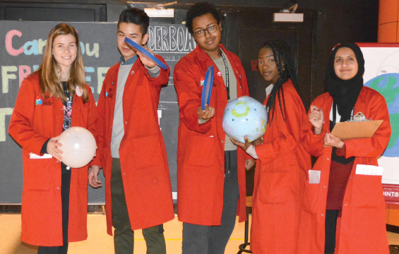 Five science school students wearing red lab coats holding a balloon, frisbees, an inflatable globe and a clipboard.