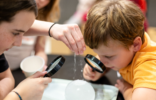 [Two kids looking through a magnifying glass at a science experiment.][Deux enfants observent une expérience scientifique à la loupe.]