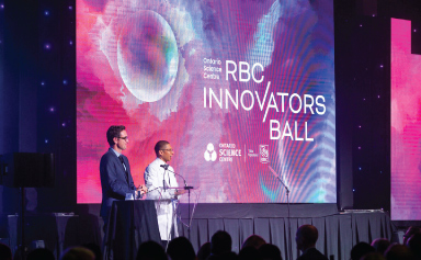 The two hosts of the ball speak at a podium on stage in front of a large screen.