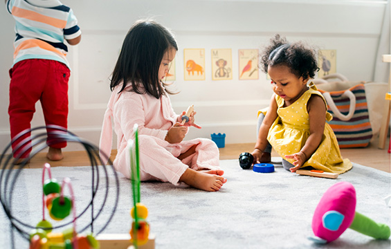 Two toddlers play with toys on a mat on the floor.