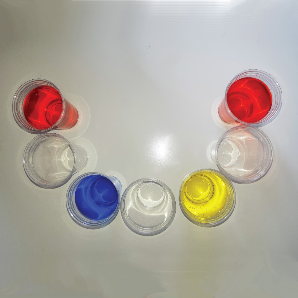 Seven plastic cups arranged in a half circle. The end two cups contain water with red colouring. The third cup contains water with blue food colouring and the fifth cup contains yellow food colouring. The second, fourth and sixth cups contain water.