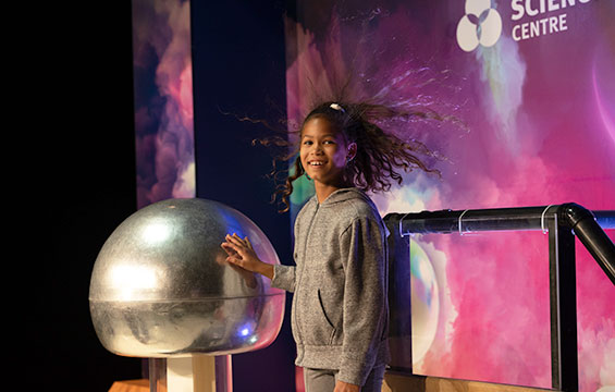 A girl holds her hand on the Van de Graaff generator while her hair stands on end.