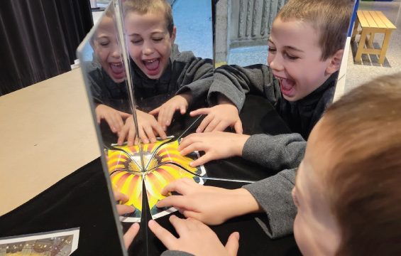 A child moves a fragment of a photo of a flower into position in front of two mirrors to create a whole image of a flower.