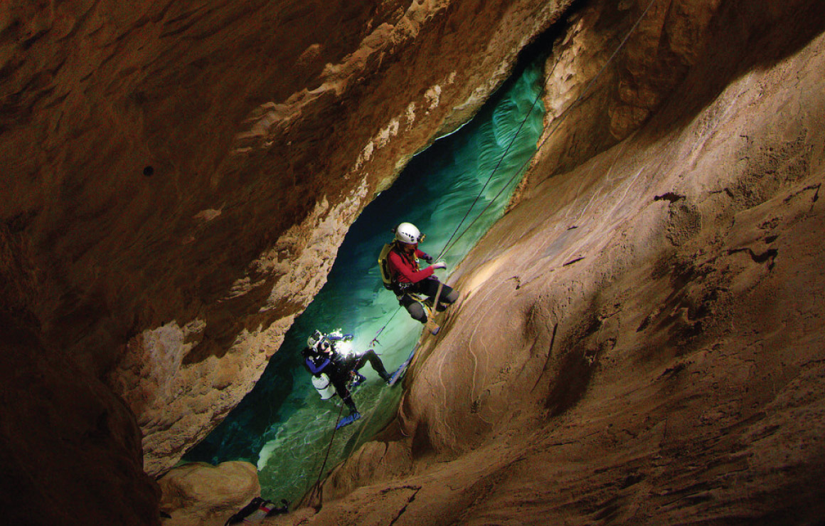 Two scientists climb up a rock wall to exit the water in a cave.