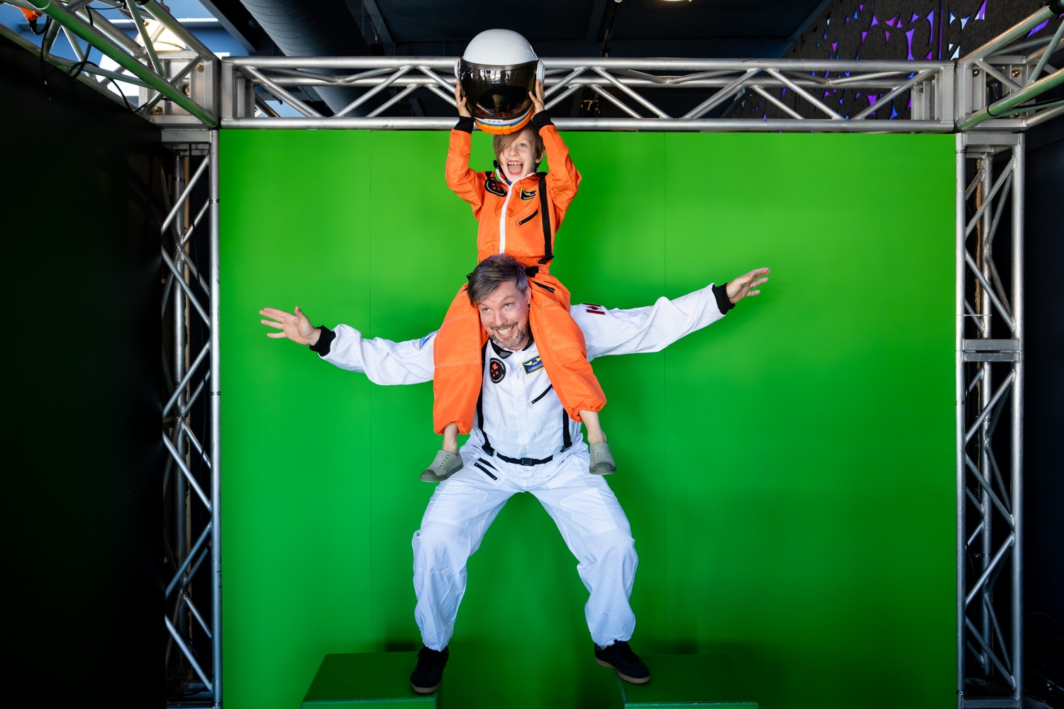 [A father and his son wearing spacesuits posing in front of the green screen.][Un père et son fils en combinaisons spatiales posent devant le fond vert.]