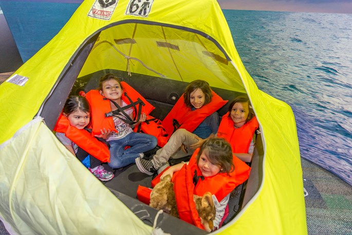 [5 kids in life jackets sit in a raft.][5 enfants en gilets de sauvetage assis dans un radeau gonflable.]