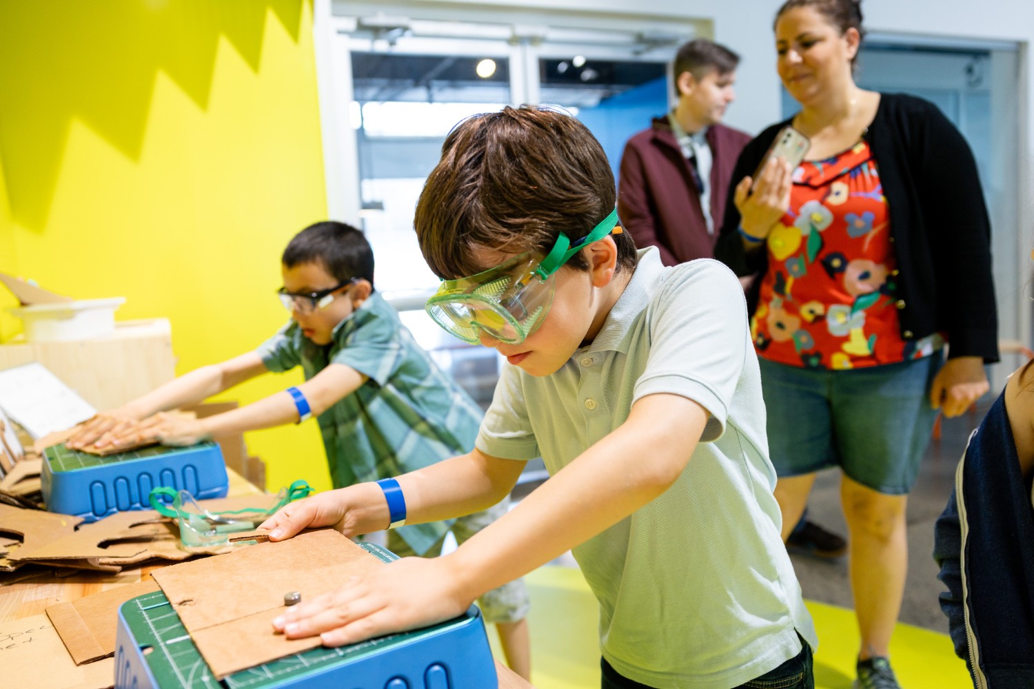 [Two boys cutting cardboards with Chomp Saw machines wearing eyeglasses to protect their eyes for safety while their parents look over their shoulders.][Deux garçons découpent des cartons avec des scies Chomp Saw, portant des lunettes de protection pour des raisons de sécurité, tandis que leurs parents les regardent par-dessus leurs épaules.]
