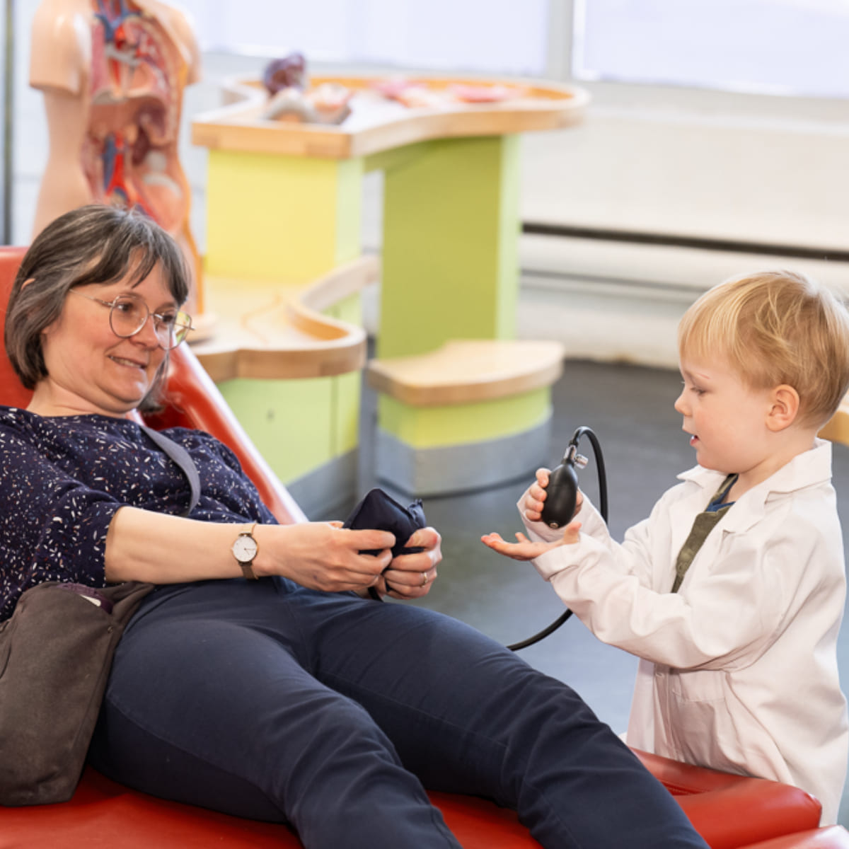 A boy wearing a doctor's coat cheecking blood pressure of his grandmother on bed.