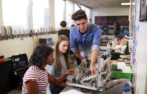 Teacher With Two Female Students Building Machine In Science Class.