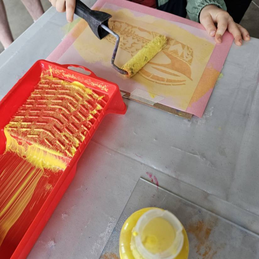 A child's hand applies a roll of yellow paint on a stencil.